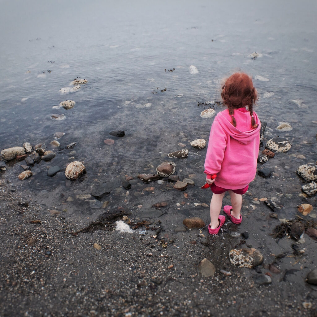 A small child in a bright pink hoodie stands at the rocky edge of a shoreline, gazing out over the calm gray water on an overcast day.