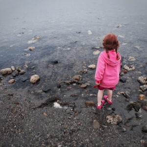 A small child in a bright pink hoodie stands at the rocky edge of a shoreline, gazing out over the calm gray water on an overcast day.