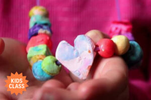 A child holds a handmade beaded necklace with bright, multicolored clay beads and a heart-shaped centerpiece.