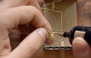 Close-up of hands assembling a small kinetic sculpture with wire, cardboard, and glue.