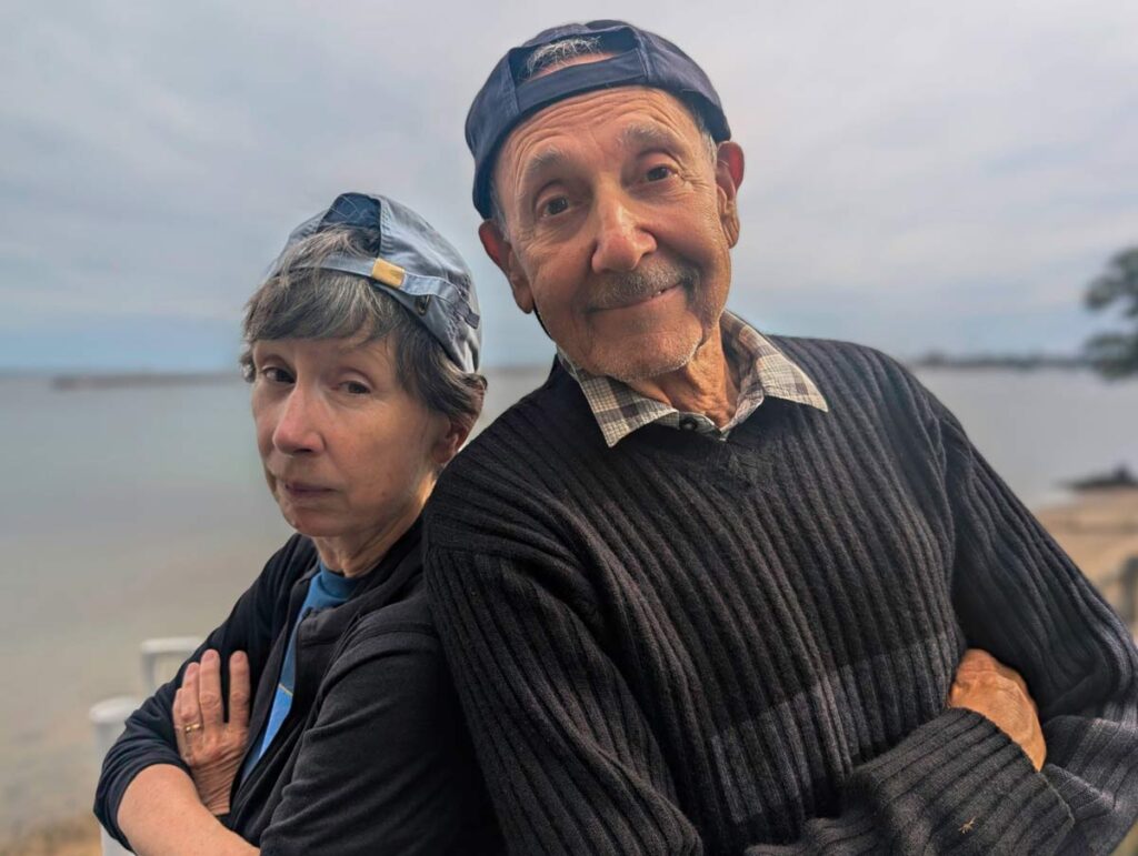 Two people standing outdoors back-to-back with arms crossed, wearing caps and smiling playfully.