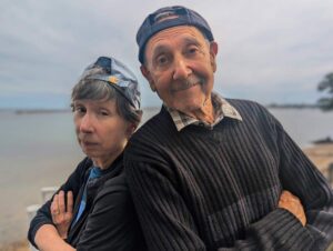 Two people standing outdoors back-to-back with arms crossed, wearing caps and smiling playfully.