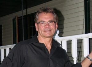 Man wearing glasses and a black shirt, standing on a porch railing.