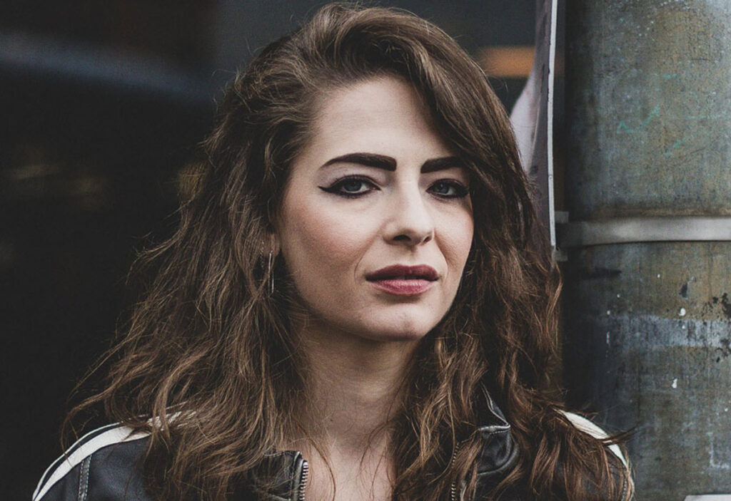 A woman with long brown hair and strong, expressive eyebrows stands in an urban outdoor setting. She wears a black leather jacket and hoop earrings, with bold eyeliner and a neutral expression. The background is softly blurred with hints of signage and metal structures.