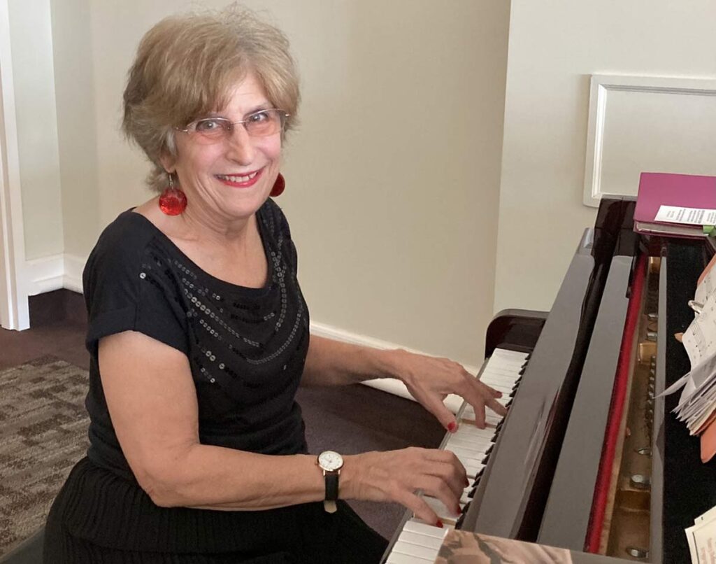 Smiling older woman with short light hair and red earrings playing the piano in a warmly lit room.