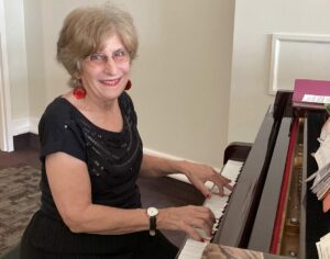 Smiling older woman with short light hair and red earrings playing the piano in a warmly lit room.
