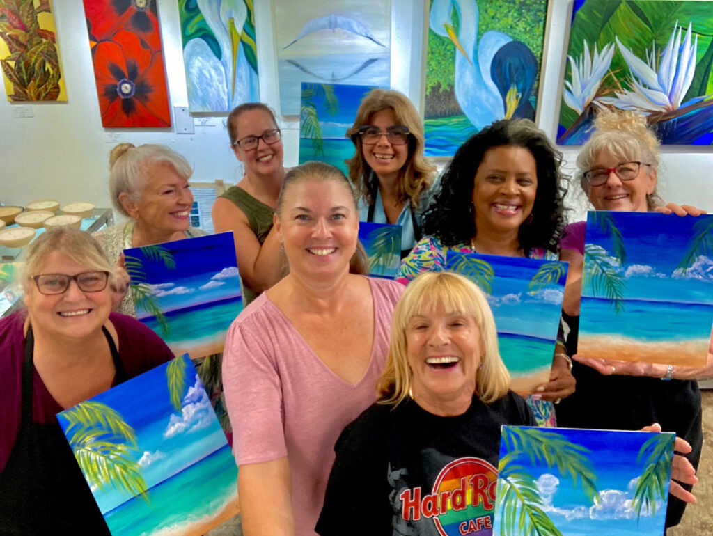 Group of smiling adults in a bright art studio holding tropical beach paintings they created, surrounded by colorful artwork on the walls.