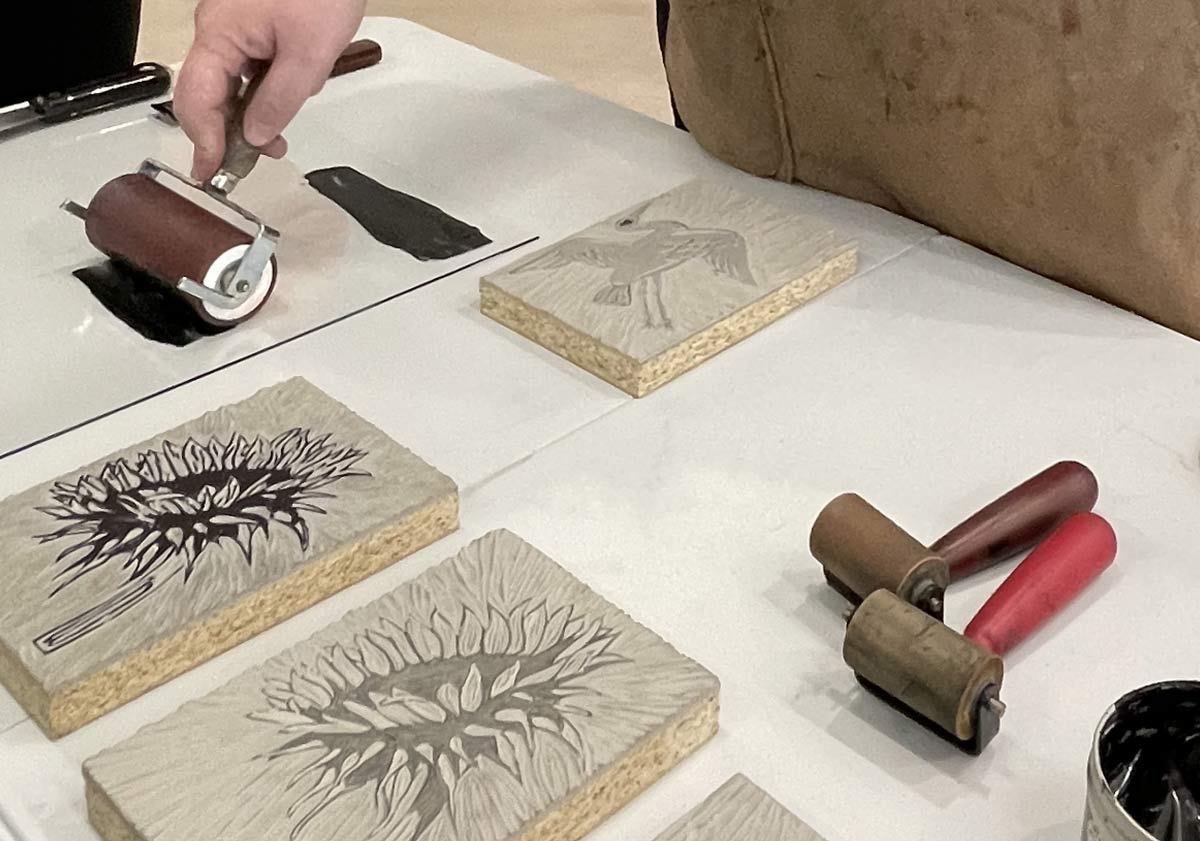 Hands rolling black ink onto carved linoleum blocks featuring floral designs.