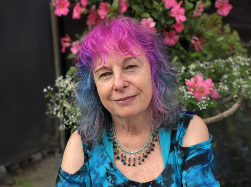 Portrait of a person with pink and blue hair sitting outdoors, smiling gently with blooming pink flowers in the background.