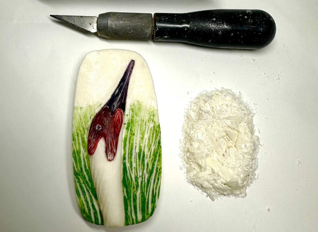 Soap carving of a bird’s head among tall grass beside carving tools.