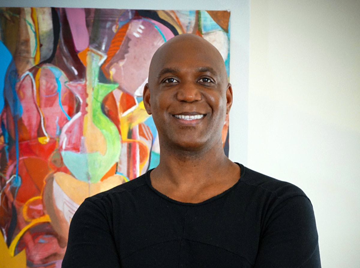 wendell-smith A man stands smiling with arms loosely crossed in front of a large, colorful abstract painting. He wears a black long-sleeve shirt. The background artwork features bold shapes in reds, blues, and greens. He appears warm, confident, and approachable.
