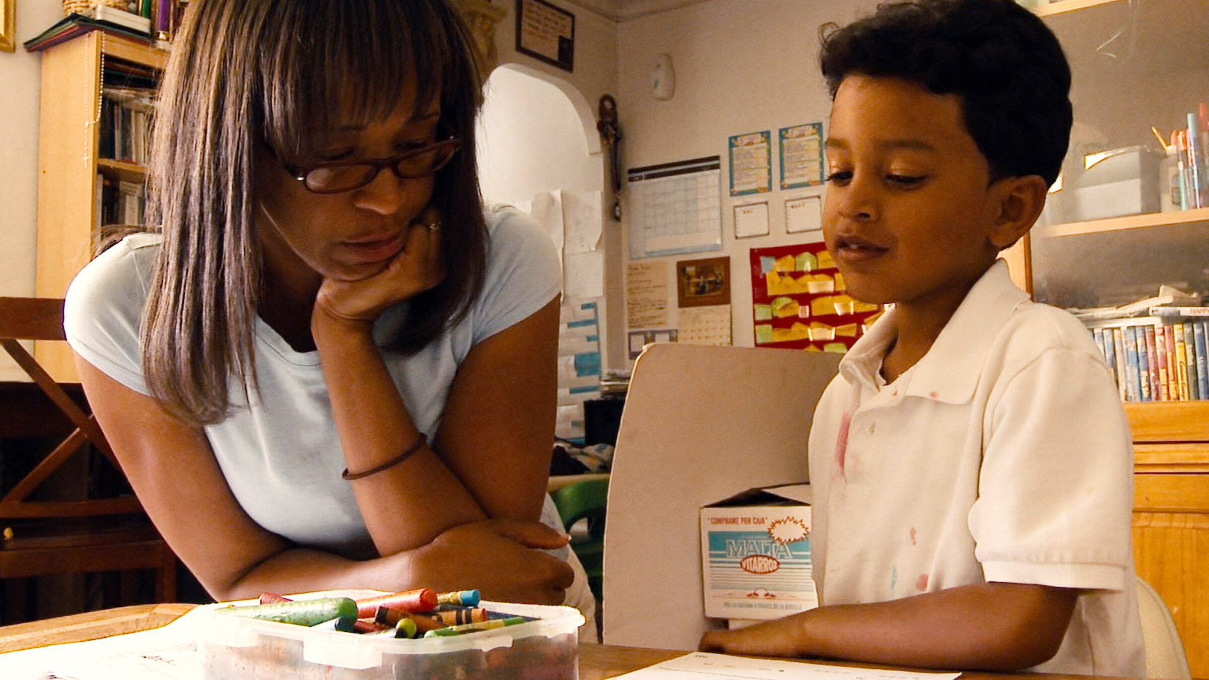 A woman sits at a table with a young boy, watching as he works with crayons and paper in a classroom or home setting.
