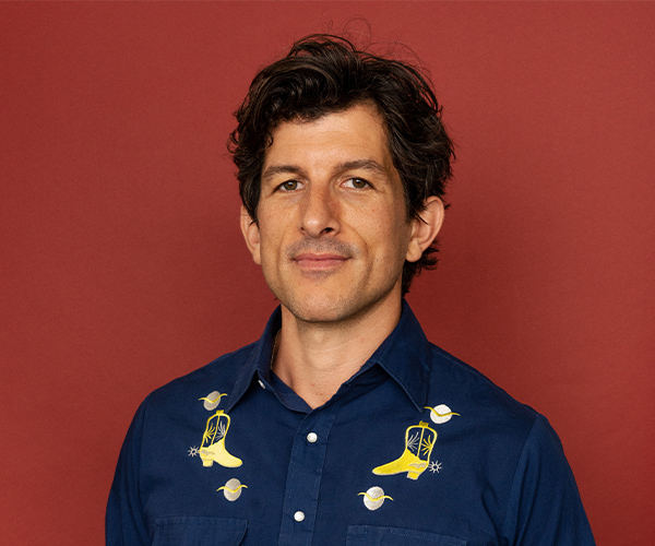 Studio headshot of a man with short dark hair, wearing a blue shirt with yellow embroidered designs, set against a red background.