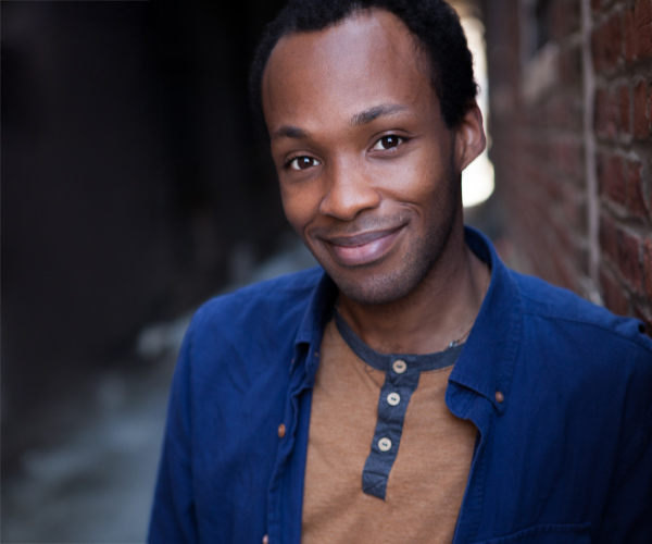 Headshot of a man with short hair, wearing a blue jacket over a brown shirt, smiling slightly in an outdoor alley setting.