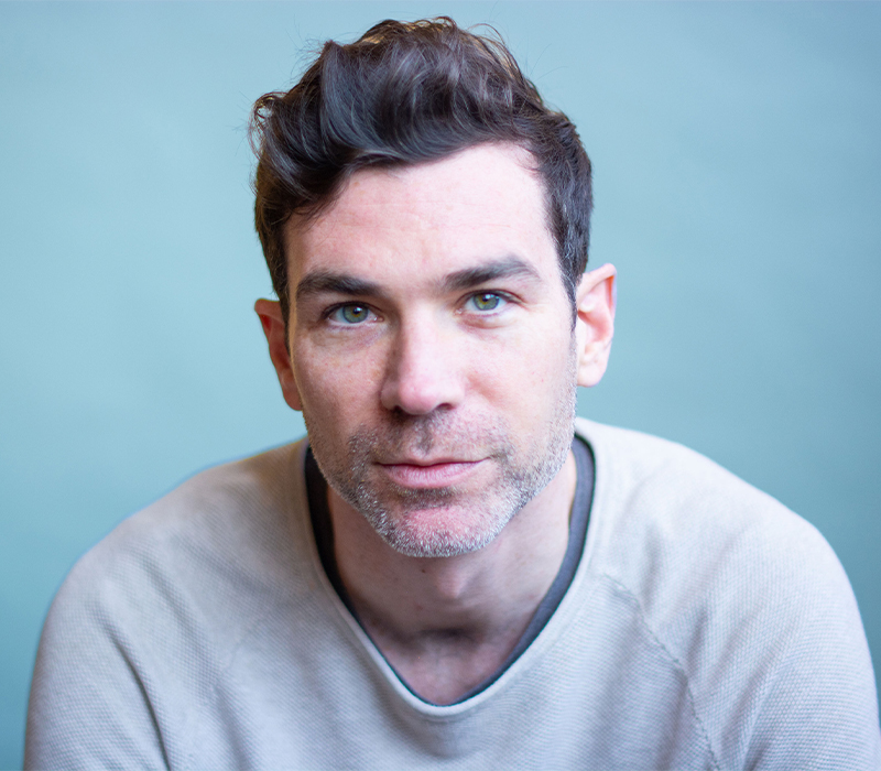 Headshot of a man with short dark hair and light stubble, wearing a light gray shirt, looking directly at the camera against a soft blue background.