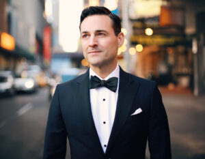 Portrait of performer Max von Essen wearing a black tuxedo and bow tie, standing on a softly lit city street with blurred lights in the background.
