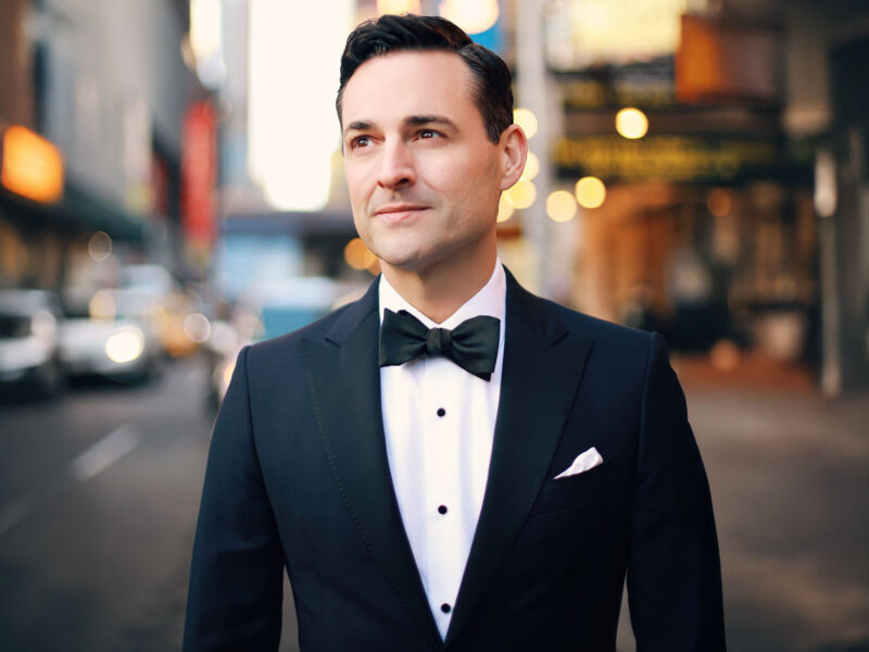 Portrait of performer Max von Essen wearing a black tuxedo and bow tie, standing on a softly lit city street with blurred lights in the background.