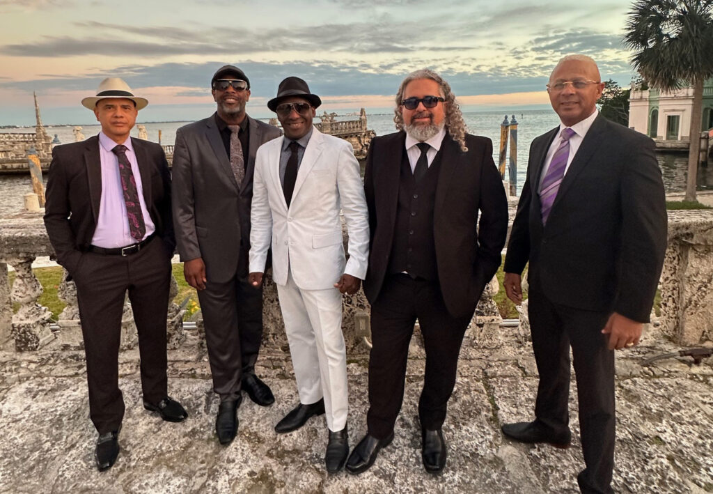 ive members of the band Cortadito stand outdoors in suits by the water at sunset, posing together with historic stone architecture behind them.