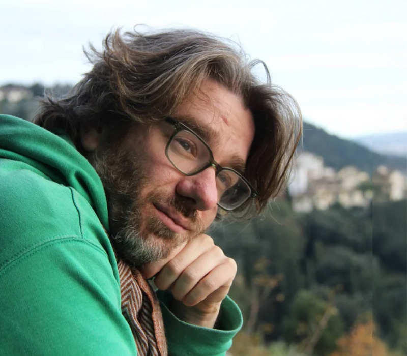 Headshot of a man with medium-length brown hair, glasses, and light facial hair, resting his chin on his hand, wearing a green hoodie with a blurred hillside landscape in the background.