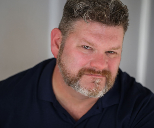 Headshot of a man with short curly hair and a trimmed beard, wearing a dark polo shirt, against a softly lit neutral background.