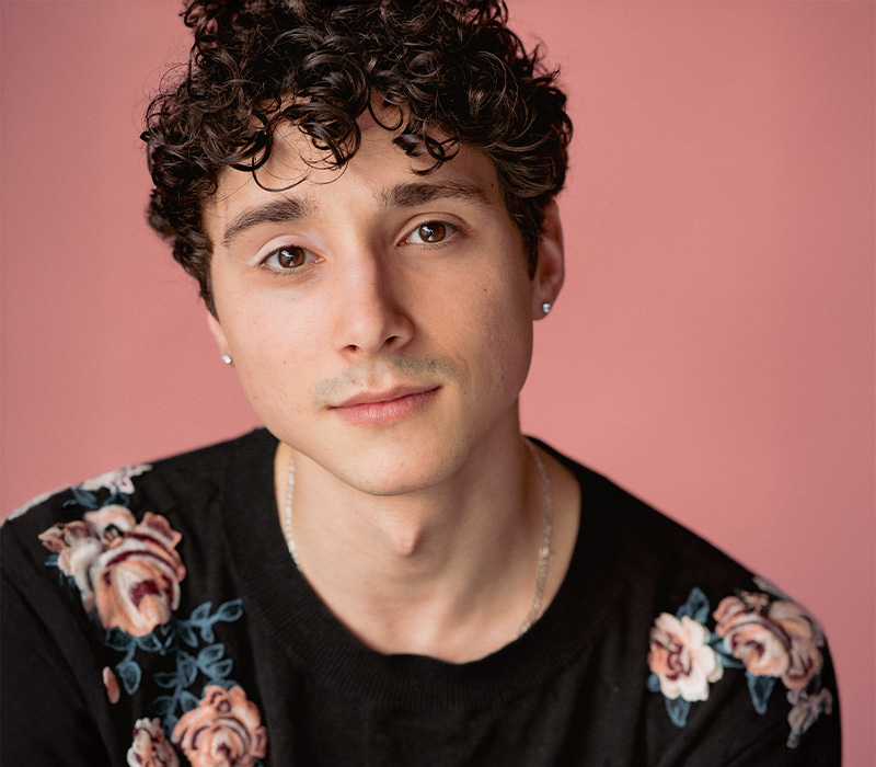 HaganOliveras Headshot of Hagan Oliveras with short curly hair, photographed against a pink background.