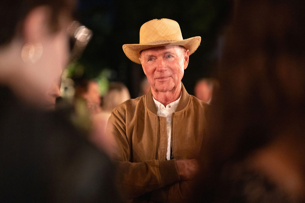 Portrait of Ed DeMore wearing a tan jacket and straw hat, standing outdoors and smiling gently while speaking with others.