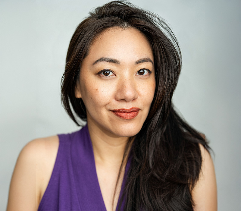 Headshot of an Asian woman with long dark hair over one shoulder, wearing a sleeveless purple top, photographed against a neutral gray background.