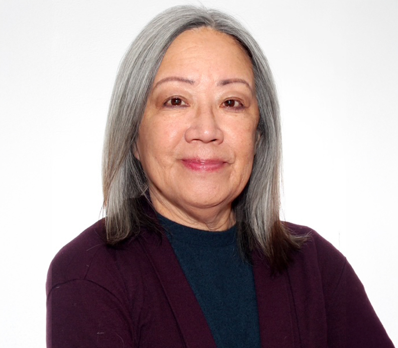 Studio headshot of an older Asian woman with straight shoulder-length gray hair, wearing a dark top and maroon cardigan against a light gray background.