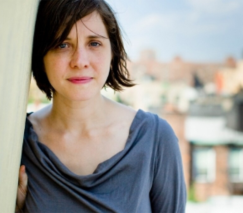 Headshot of a woman with short dark brown hair, leaning against a wall, wearing a soft gray top, with a blurred outdoor cityscape in the background.