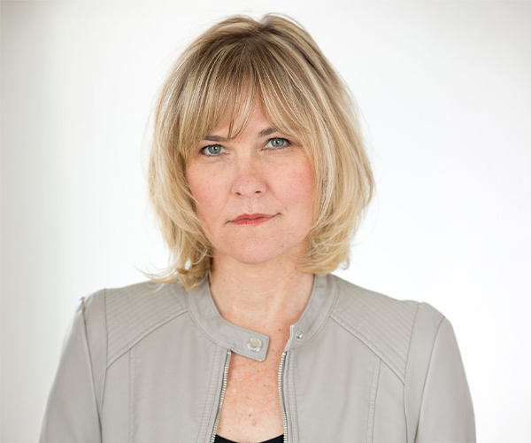 Studio headshot of a woman with a blonde bob haircut, wearing a light gray jacket, against a plain white background.
