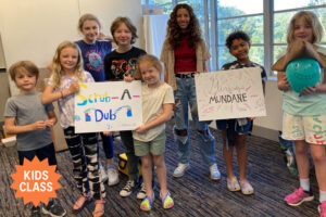 Group of smiling kids in a classroom holding handmade posters that read “Scrub-A-Dub Commercial” and “The Miraculous Mundane Show,” suggesting a creative youth theater or film project.