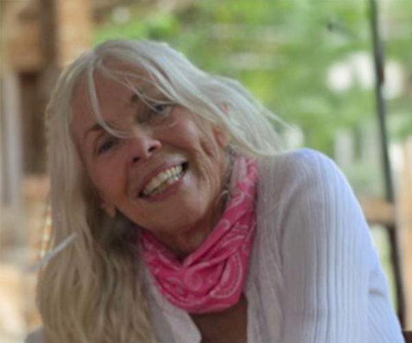 Outdoor headshot of an older woman with long white hair, smiling and wearing a pink scarf and light top, with greenery in the background.