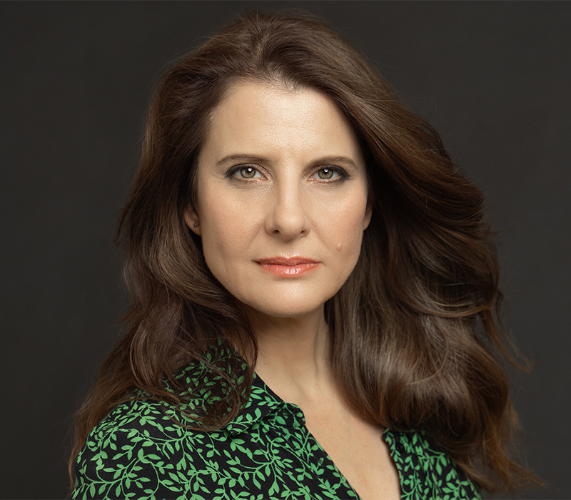 Studio headshot of a woman with long wavy brown hair, wearing a green patterned blouse, set against a dark background.