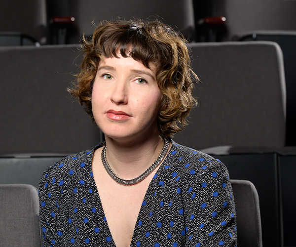 Headshot of a woman with short curly brown hair, wearing a blue polka dot blouse, seated in a theater with rows of seats behind her.