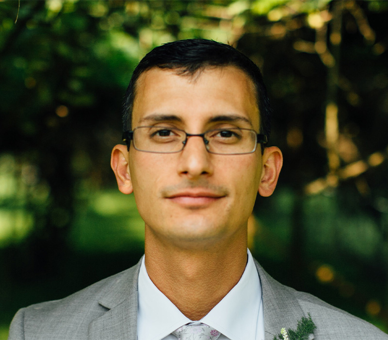 Nick Gandiello Headshot of Nick Gandiello wearing glasses and a gray suit, photographed outdoors with a soft green background.