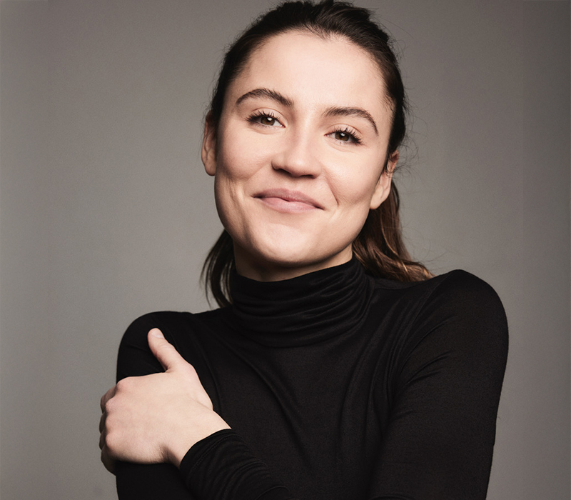 Headshot of a woman with hair pulled back, wearing a black turtleneck, smiling softly with arms crossed against a neutral gray background.