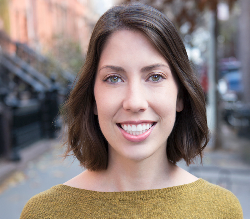 ashley-monroe Headshot of an actor with short brown hair, smiling, wearing a mustard-yellow top, photographed outdoors on a city street.