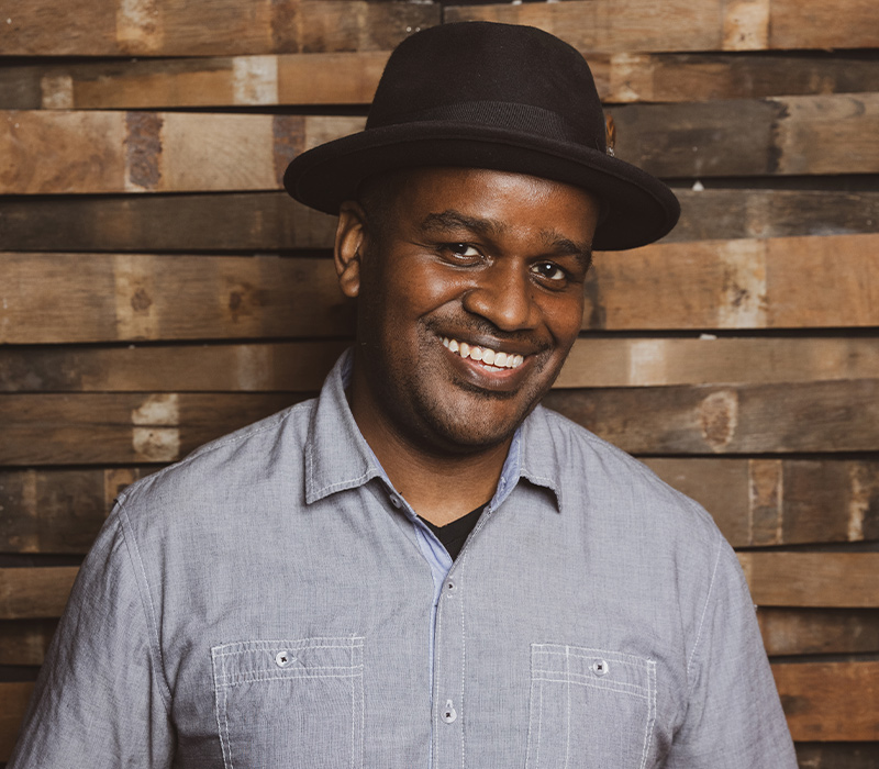 shawn-randall Headshot of an actor smiling, wearing a dark hat and light button-down shirt, standing against a wood-plank background.