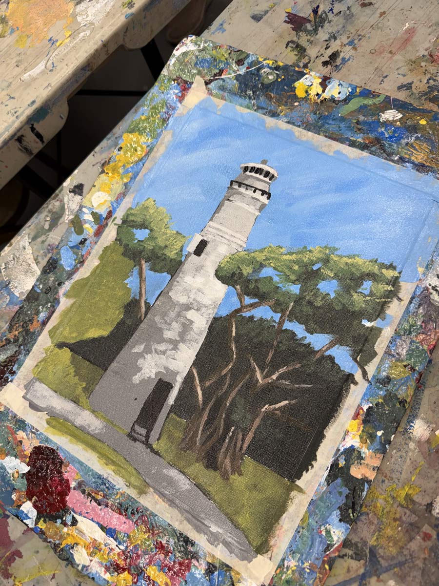 Painting of a lighthouse with trees and shadows on a work table with mixed paint.