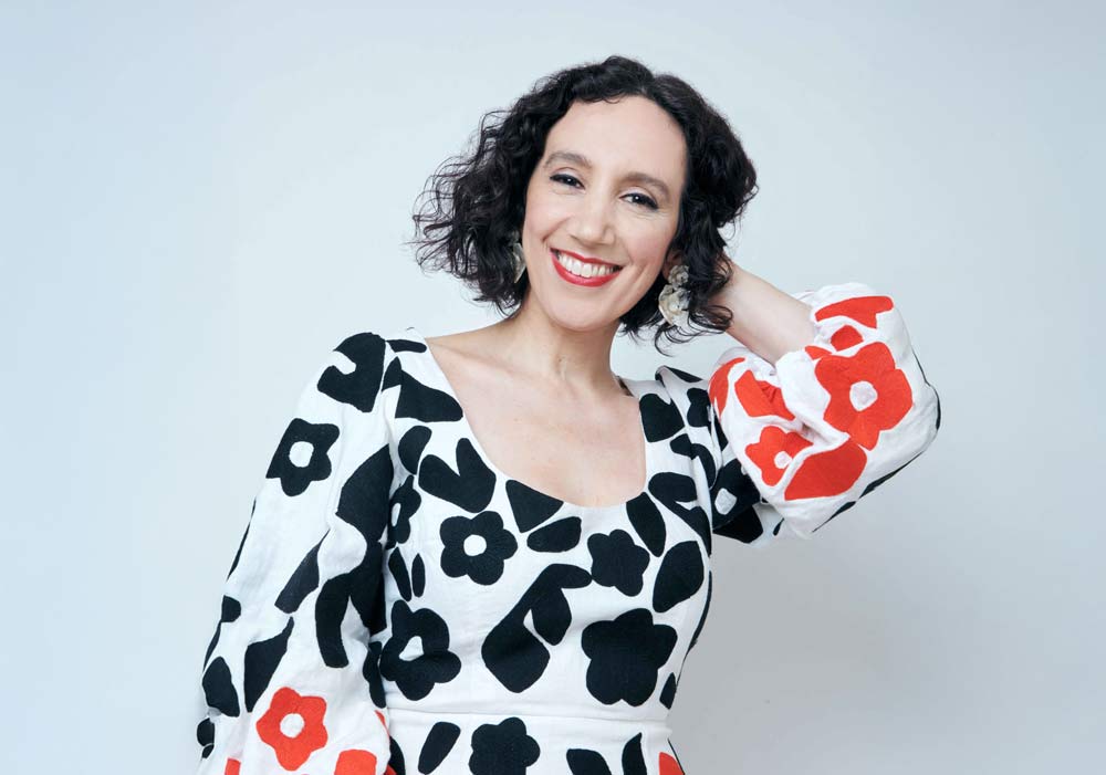 Gabrielle Singer Gabrielle Stravelli smiling in a white dress with black and red floral patterns against a light gray background.