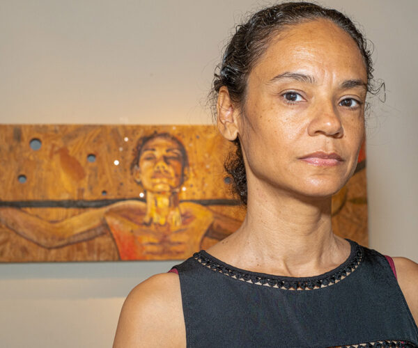 Portrait of artist Sally Binard standing indoors in front of one of her paintings, wearing a sleeveless black top and looking directly at the camera.
