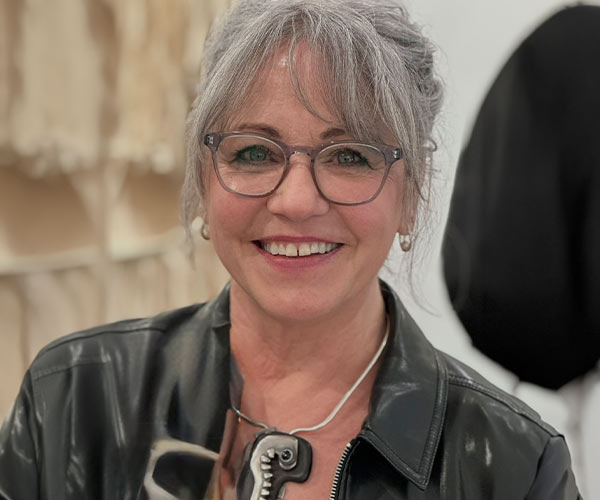 Portrait of a woman with grey hair and bangs wearing glasses and a black leather jacket standing in a studio space.