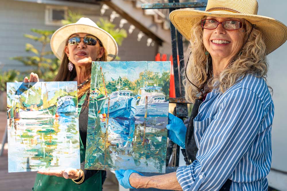 Two women outdoors holding colorful boat paintings during plein air painting class