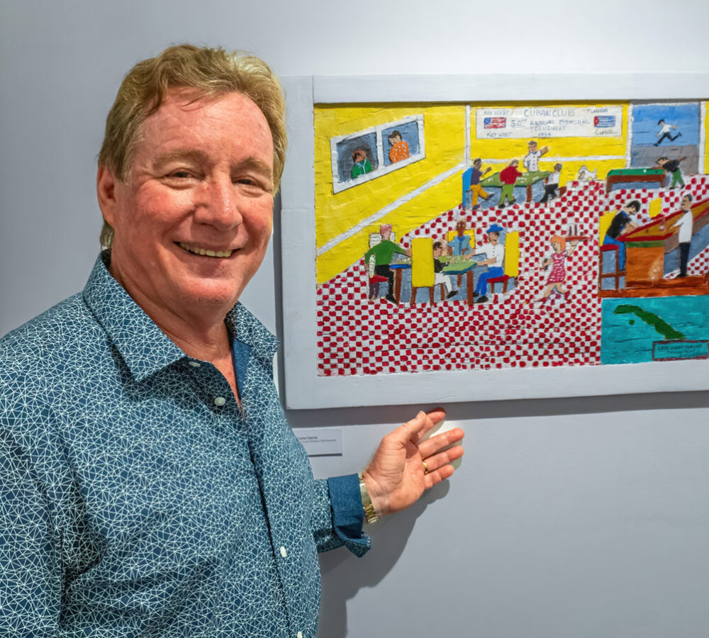 Smiling artist standing beside colorful woodcut intaglio depicting lively indoor scene