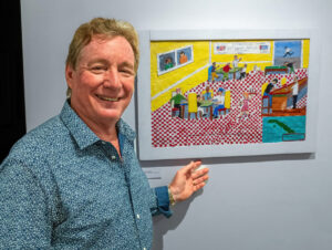Smiling artist standing beside colorful woodcut intaglio depicting lively indoor scene