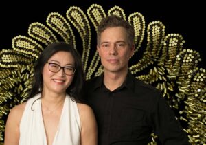 Pianist Jiayin Shen and visual artist Eric Dyer pose before a dramatic backdrop of brass shell casings arranged in a radiant pattern.